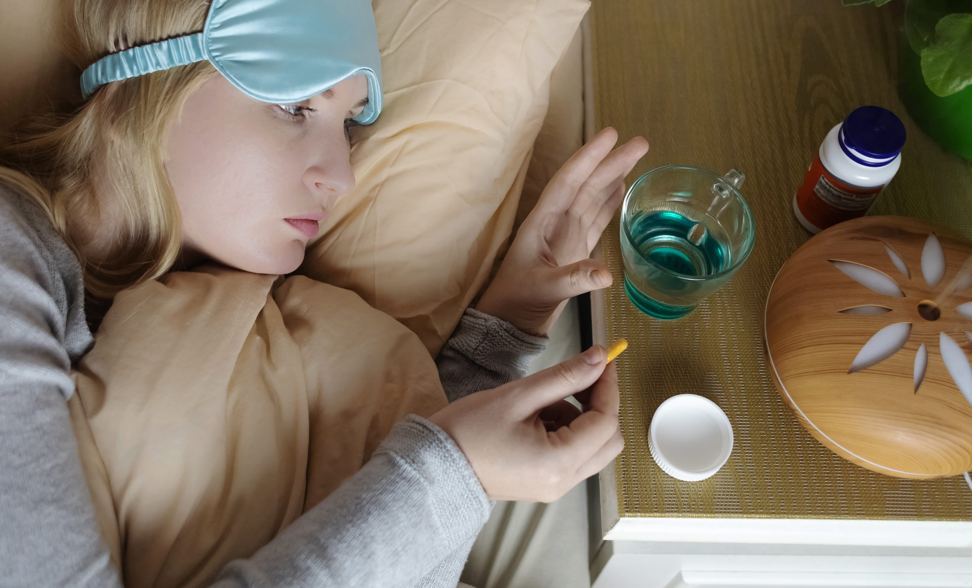 Woman wearing a sleep mask reaching for medication on her bedside table, representing taking melatonin for sleep.