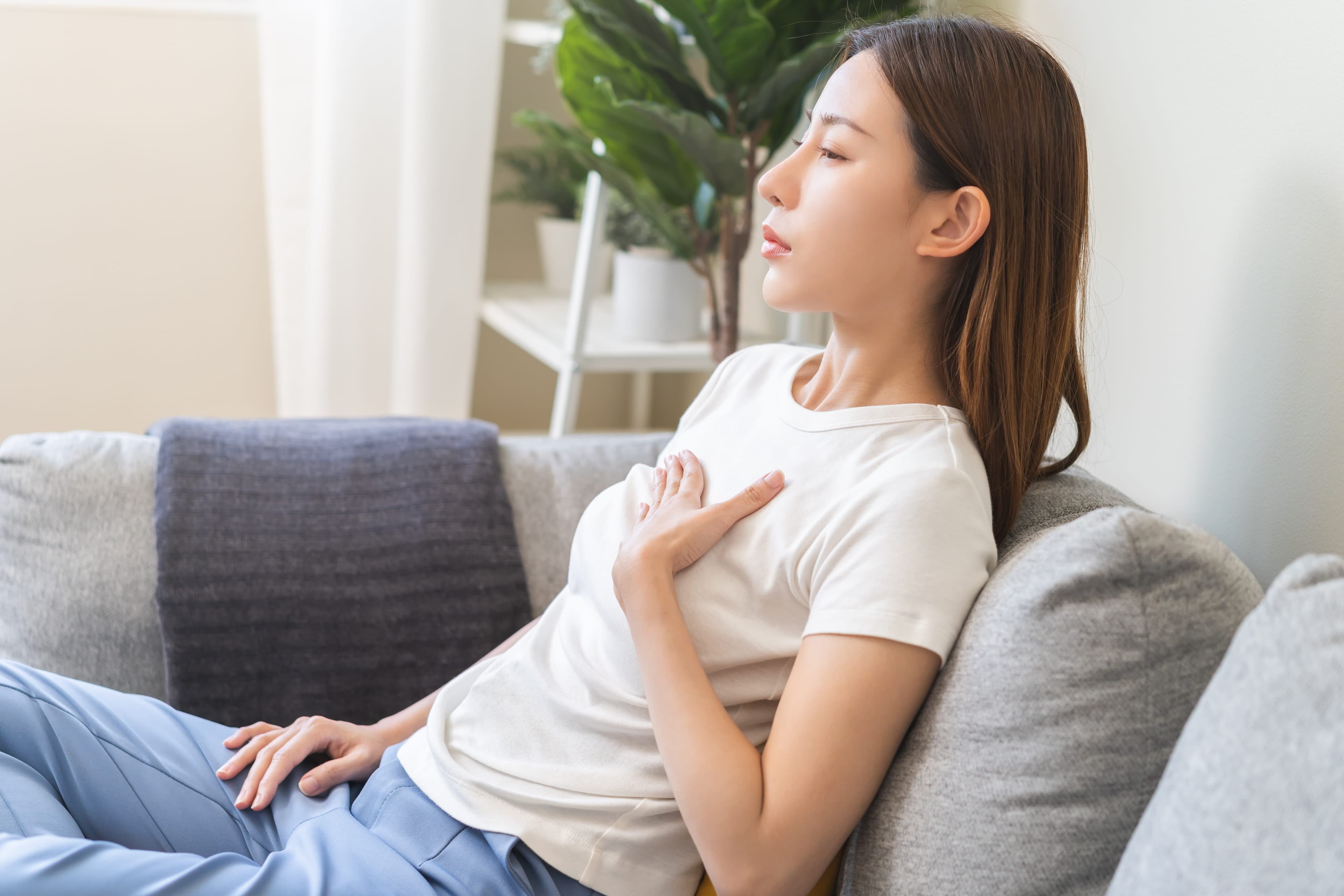 Woman sitting on her sofa with a hand on her chest, feeling the discomfort of acid reflux and heartburn.