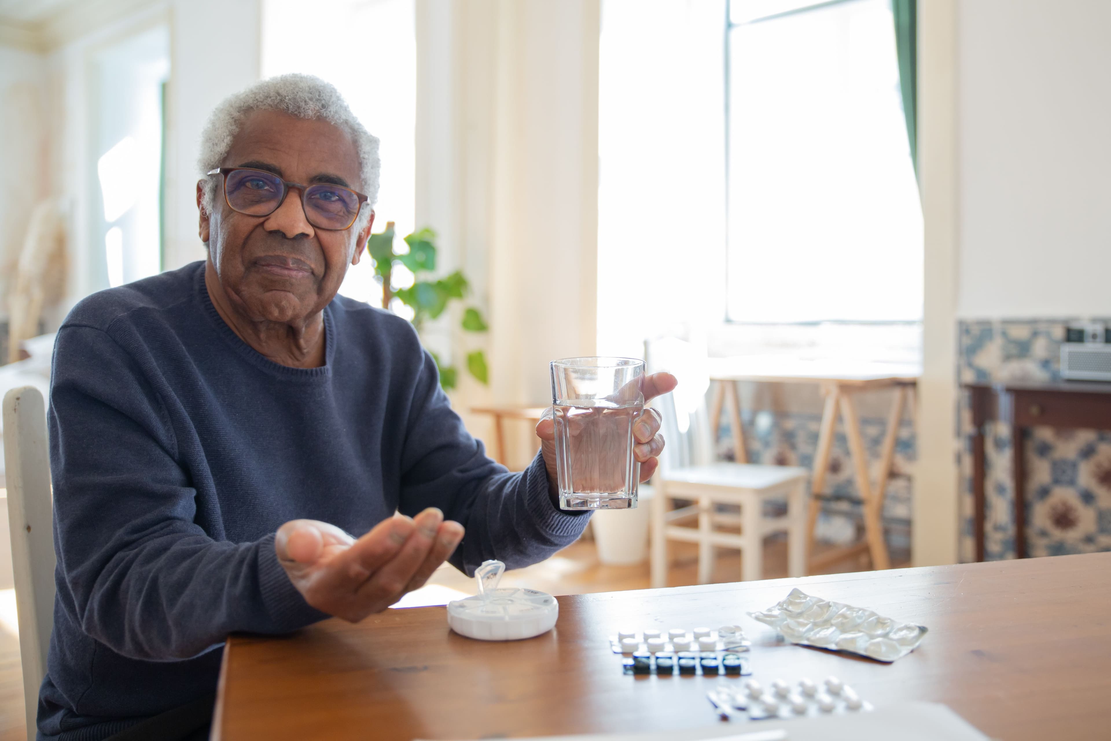 Older man holding a glass of water beside his tablets and pill organiser, taking his medication as prescribed.